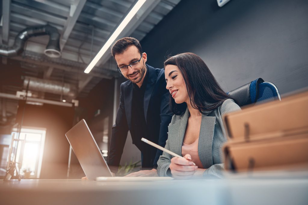 Happy young colleagues are looking at laptop screen while talking in modern office together